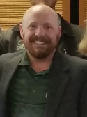 A person with light skin, a light brown beard, and no head hair is wearing a dark green button up shirt with a gray blazer on top